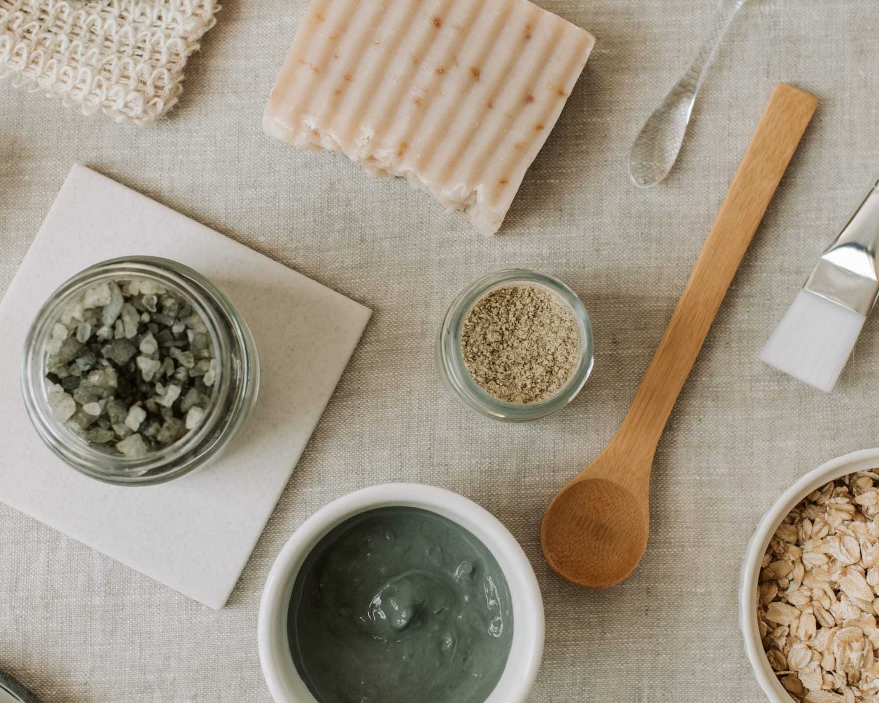 Natural spa ingredients including soap, sea salt, clay mask, oats, and a wooden spoon on fabric.