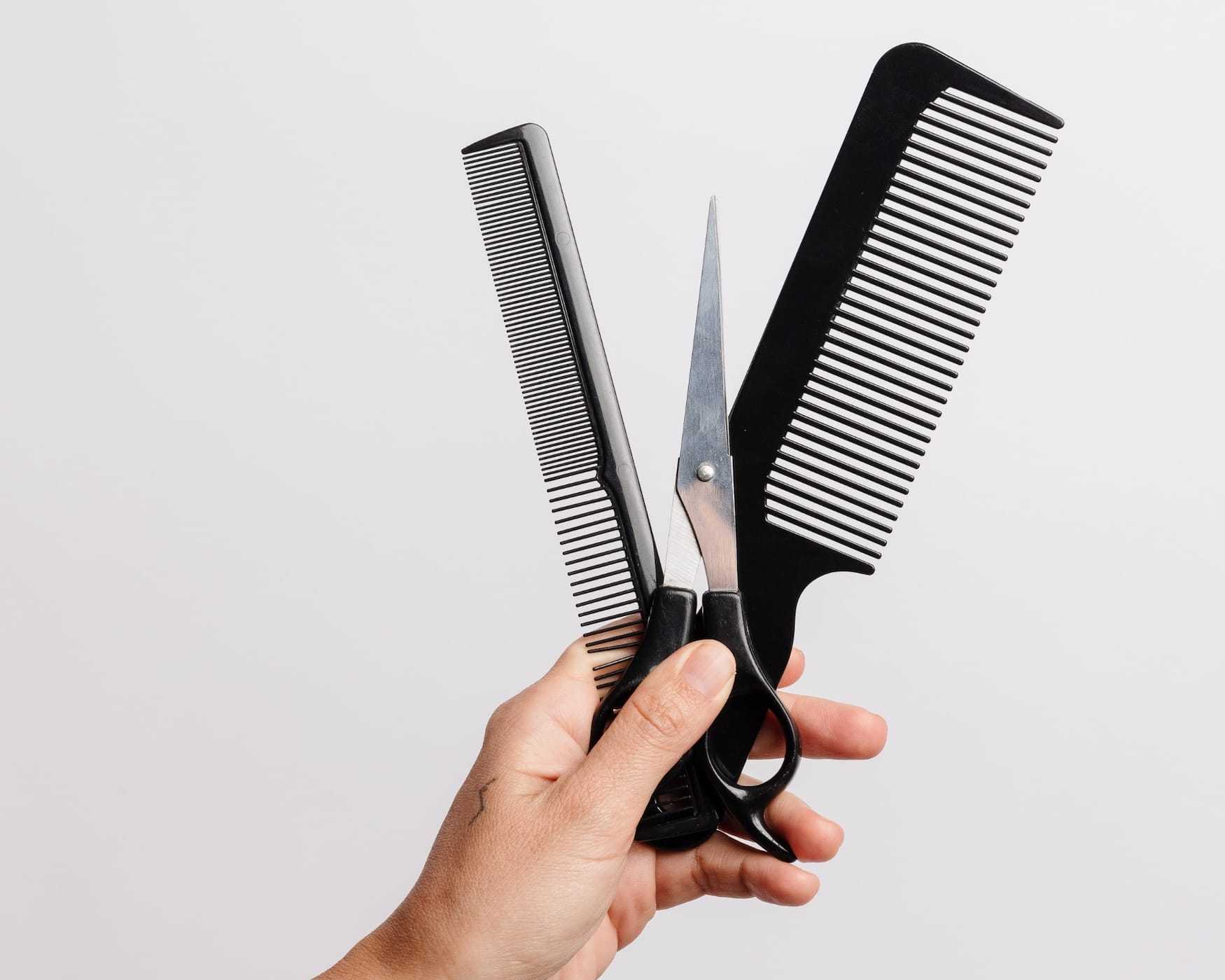 Hand holding a pair of scissors and two black hair combs against a white background.