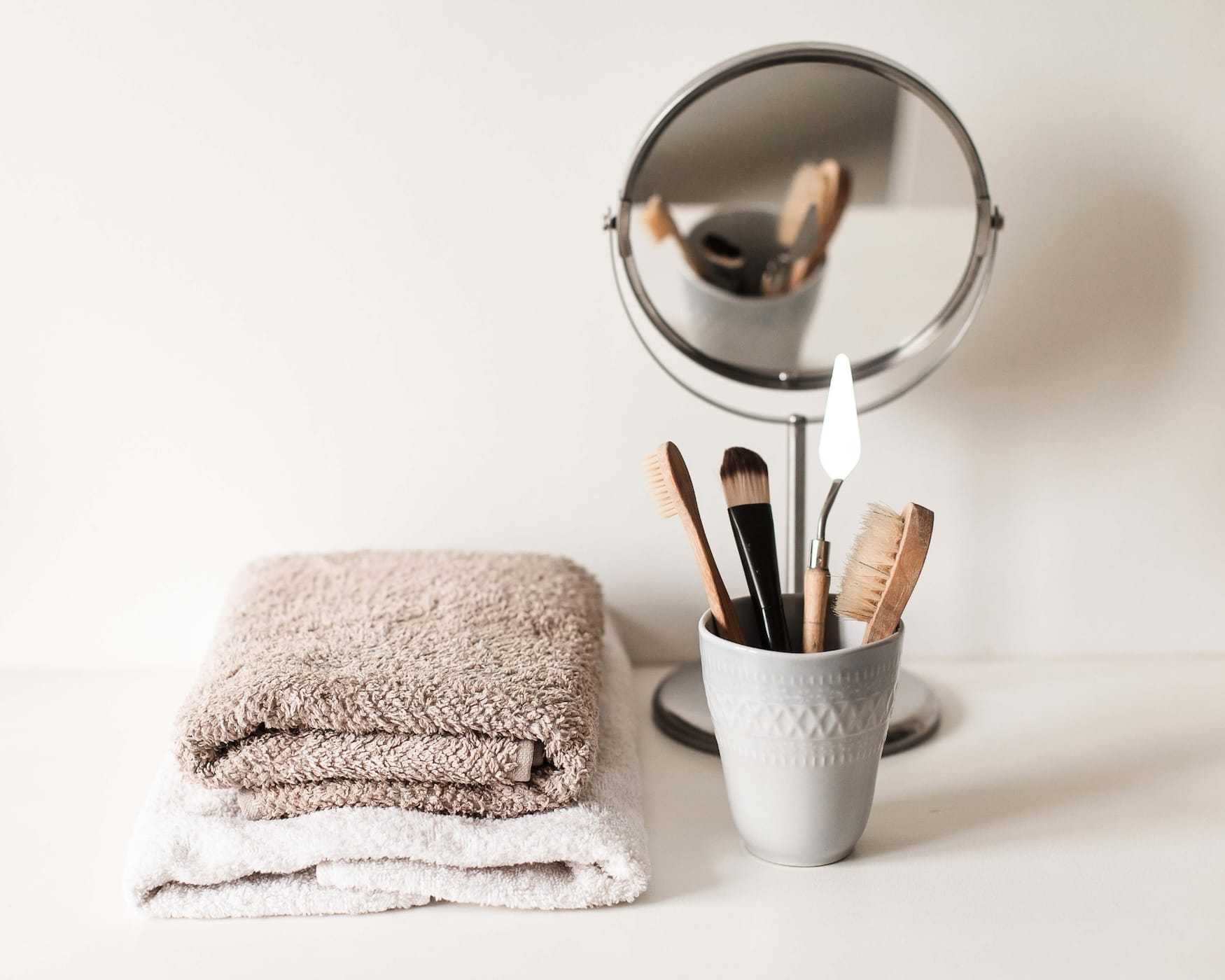 Stacked towels, vanity mirror, and toothbrushes in a white holder on a light-colored surface.